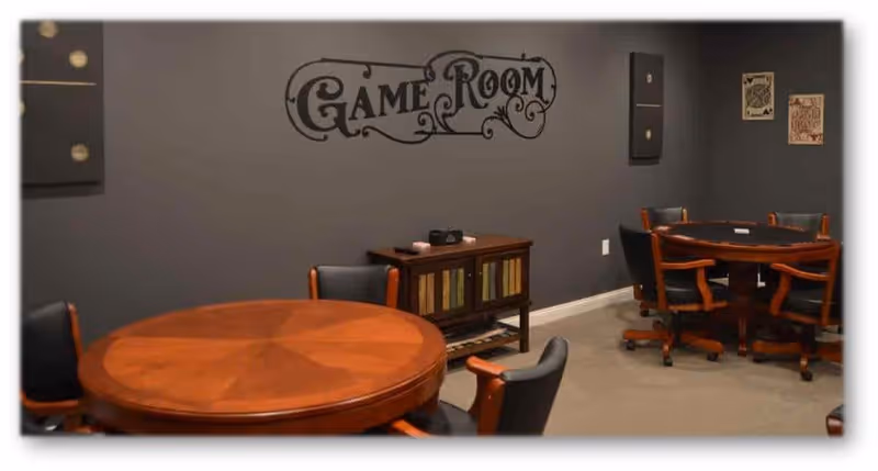 Interior view of a game room with two round wooden tables surrounded by black cushioned chairs with wooden armrests. The walls are painted gray and decorated with a 'Game Room' sign and framed pictures. A small wooden cabinet is placed against the wall between the tables.