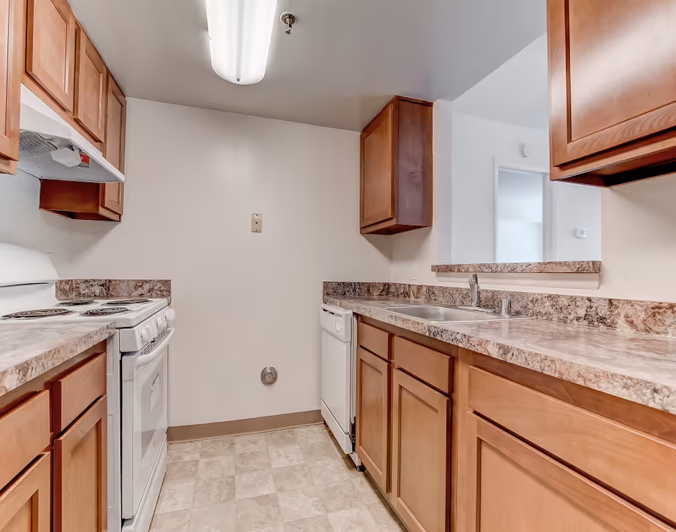 A compact kitchen with wooden cabinets, a white electric stove with oven, a white dishwasher, and a double sink with a faucet. The countertops have a marbled pattern, and the floor is covered with light-colored tiles. The walls are plain white, and there is a fluorescent ceiling light fixture.