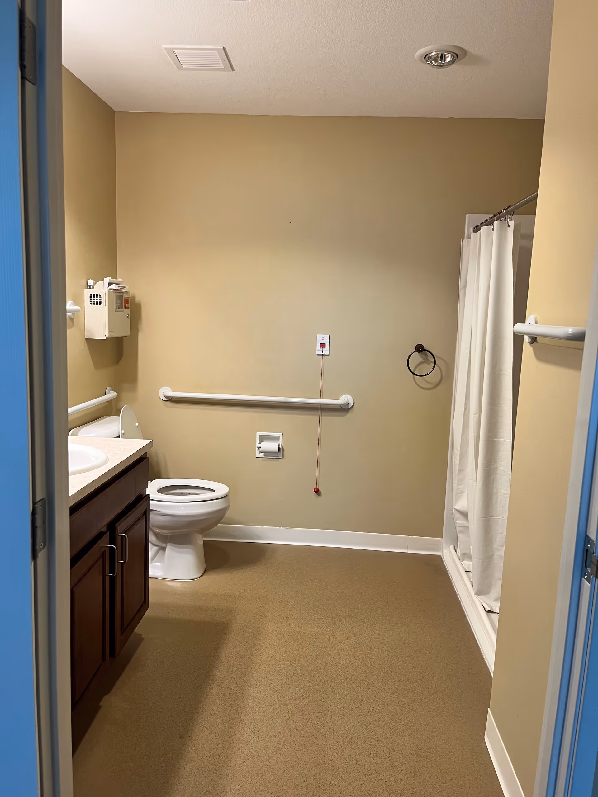 A clean and accessible bathroom with beige walls and flooring. It features a white toilet with grab bars on the wall behind and beside it, a white sink with a dark wooden cabinet, a shower with a white curtain, a towel ring, and an emergency pull cord on the wall.