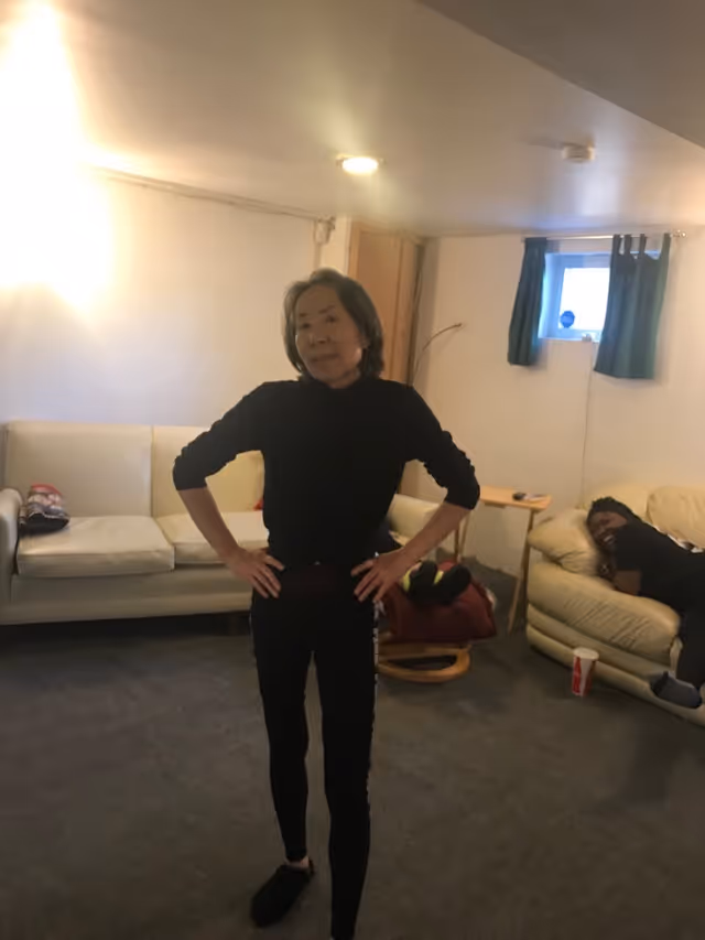 A person stands with hands on hips in a living room area with sofas and another person reclining on a couch in the background.