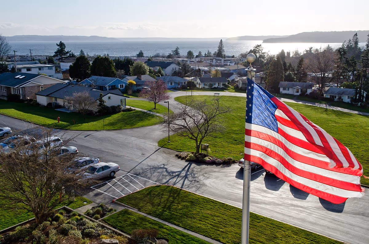 View of a residential neighborhood with houses, green lawns, and a large American flag in the foreground. In the background, there is a body of water and distant hills under a partly cloudy sky.