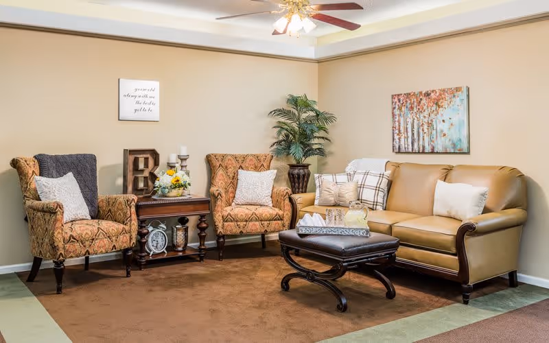 A cozy living room area with a tan leather sofa, two patterned armchairs with white pillows, a dark wooden coffee table with a tray and pitcher, a side table with decorative items, a potted plant, and wall art including a framed quote and a colorful painting of trees.