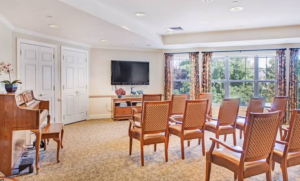 A bright room with multiple wooden chairs arranged in rows facing a wall-mounted flat screen TV. There is a wooden piano with a bench on the left side, a small wooden cabinet under the TV with decorative items, and large windows with floral curtains letting in natural light. The room has beige walls and patterned carpet flooring.