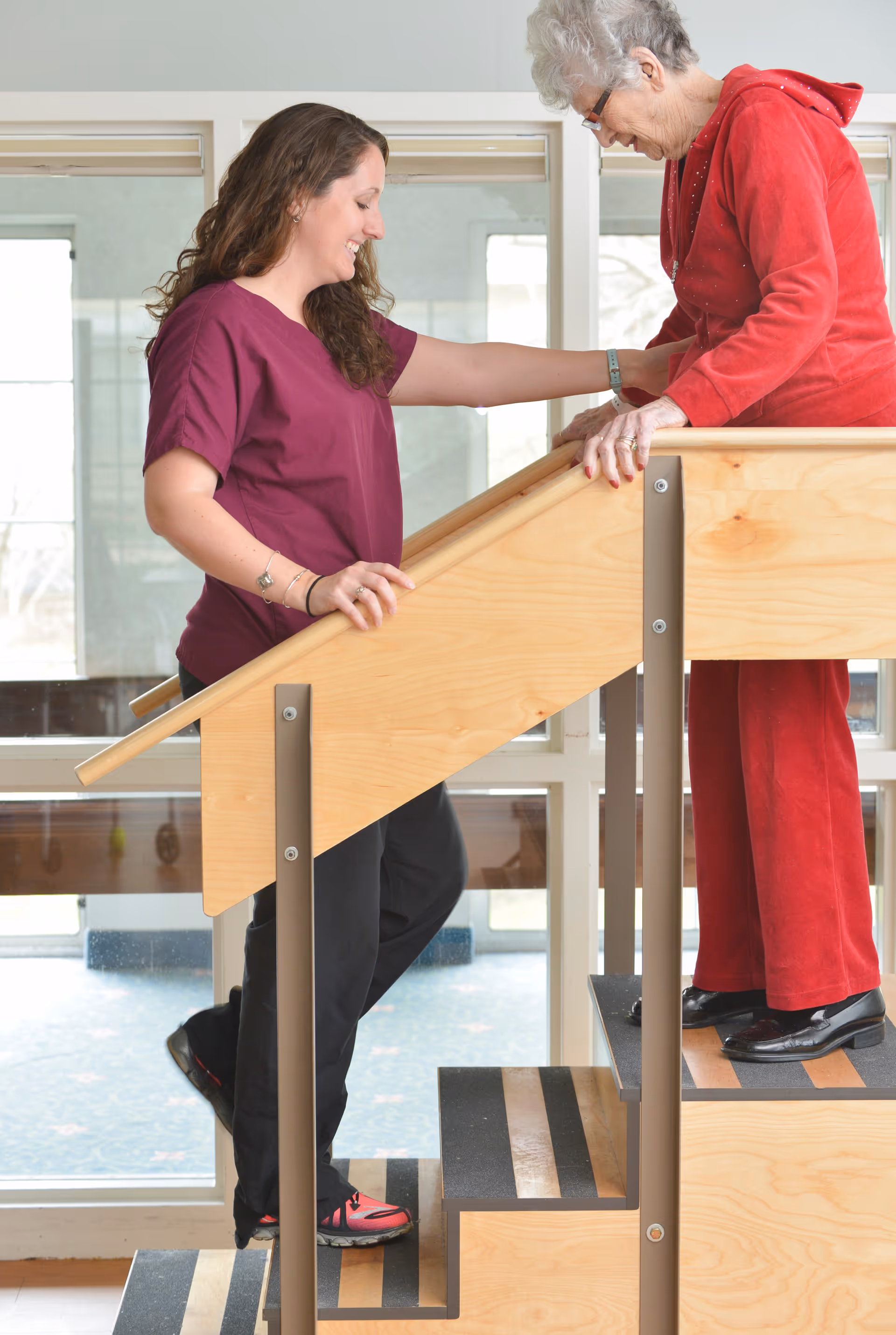 A caregiver in a maroon shirt assists an elderly woman dressed in red as she walks up a small set of wooden stairs indoors, with large windows in the background.