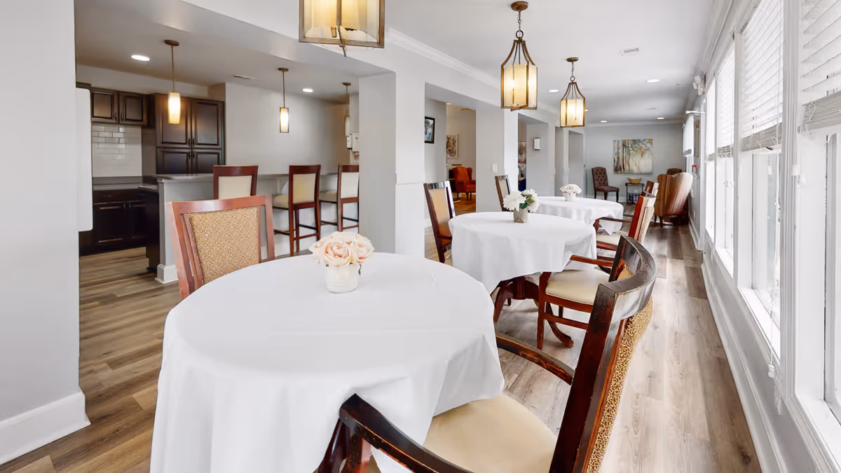 Sunlit dining area with round tables covered in white linens, wooden chairs, bar seating, and large windows along one side.