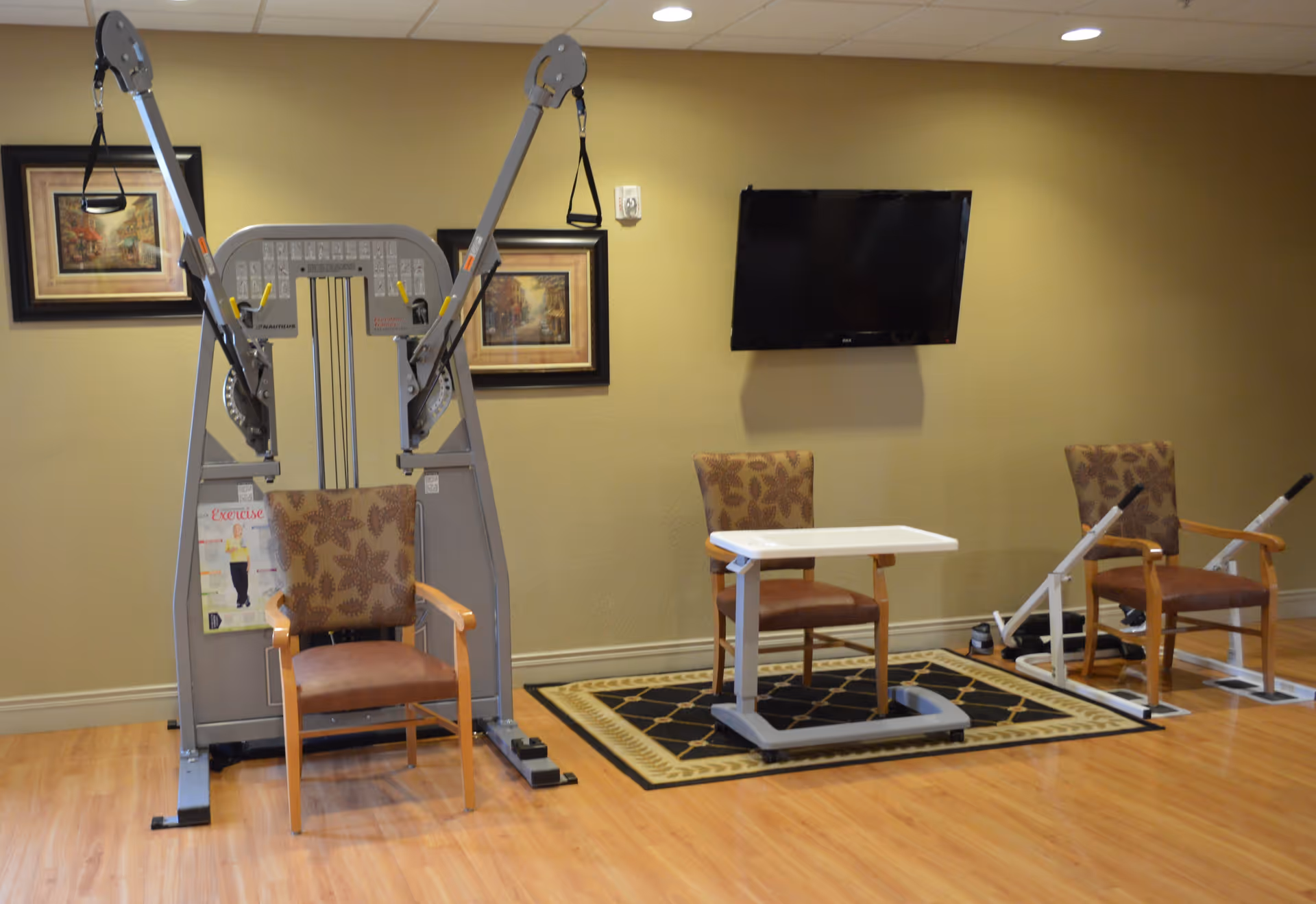 A small interior therapy/exercise room with exercise machines, three chairs, a movable table, a wall-mounted TV and framed pictures.