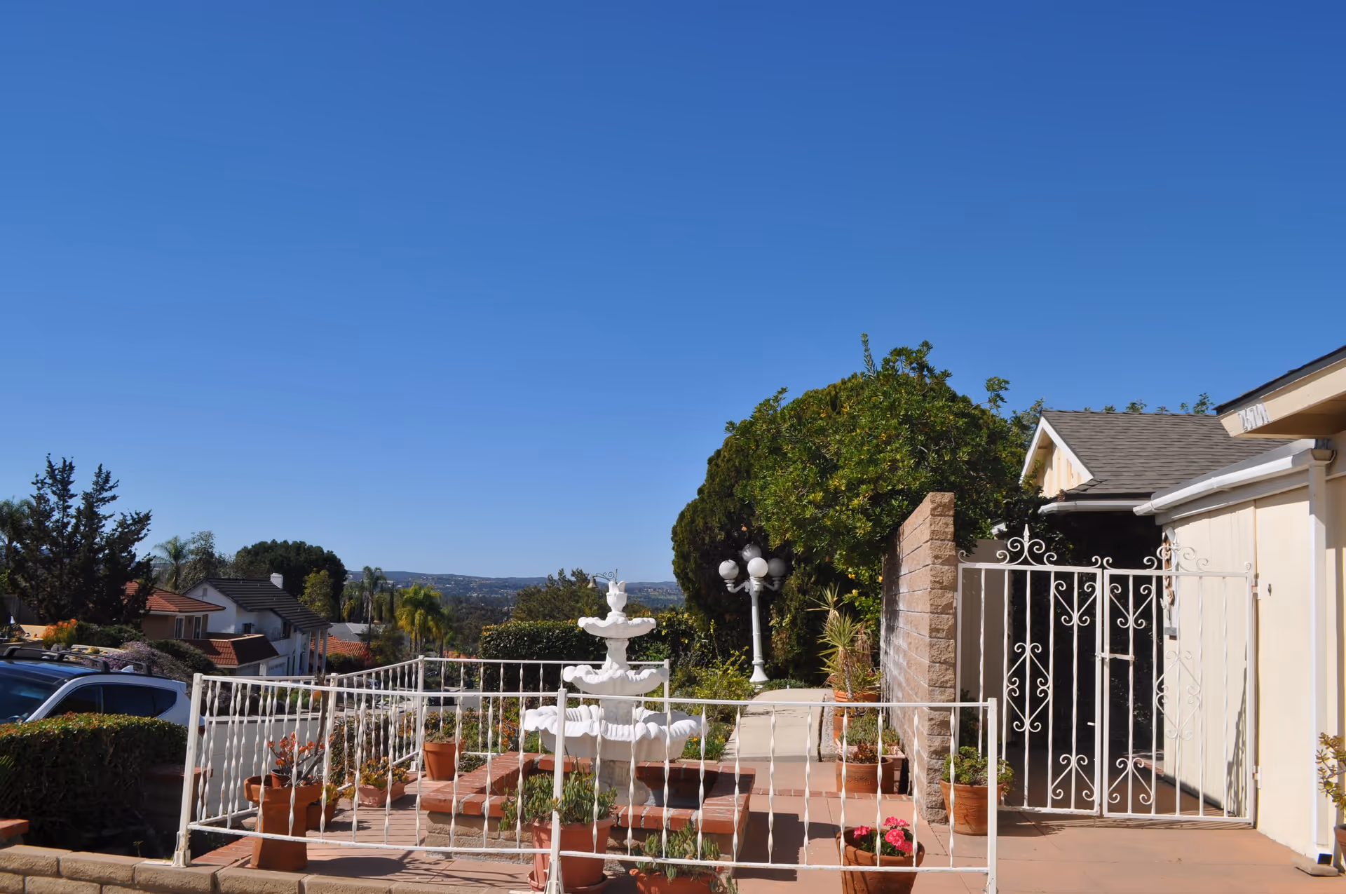 Outdoor view of a senior living facility showing a white decorative fountain surrounded by potted plants, a white metal fence and gate, a walkway, and a clear blue sky with trees and houses in the background.