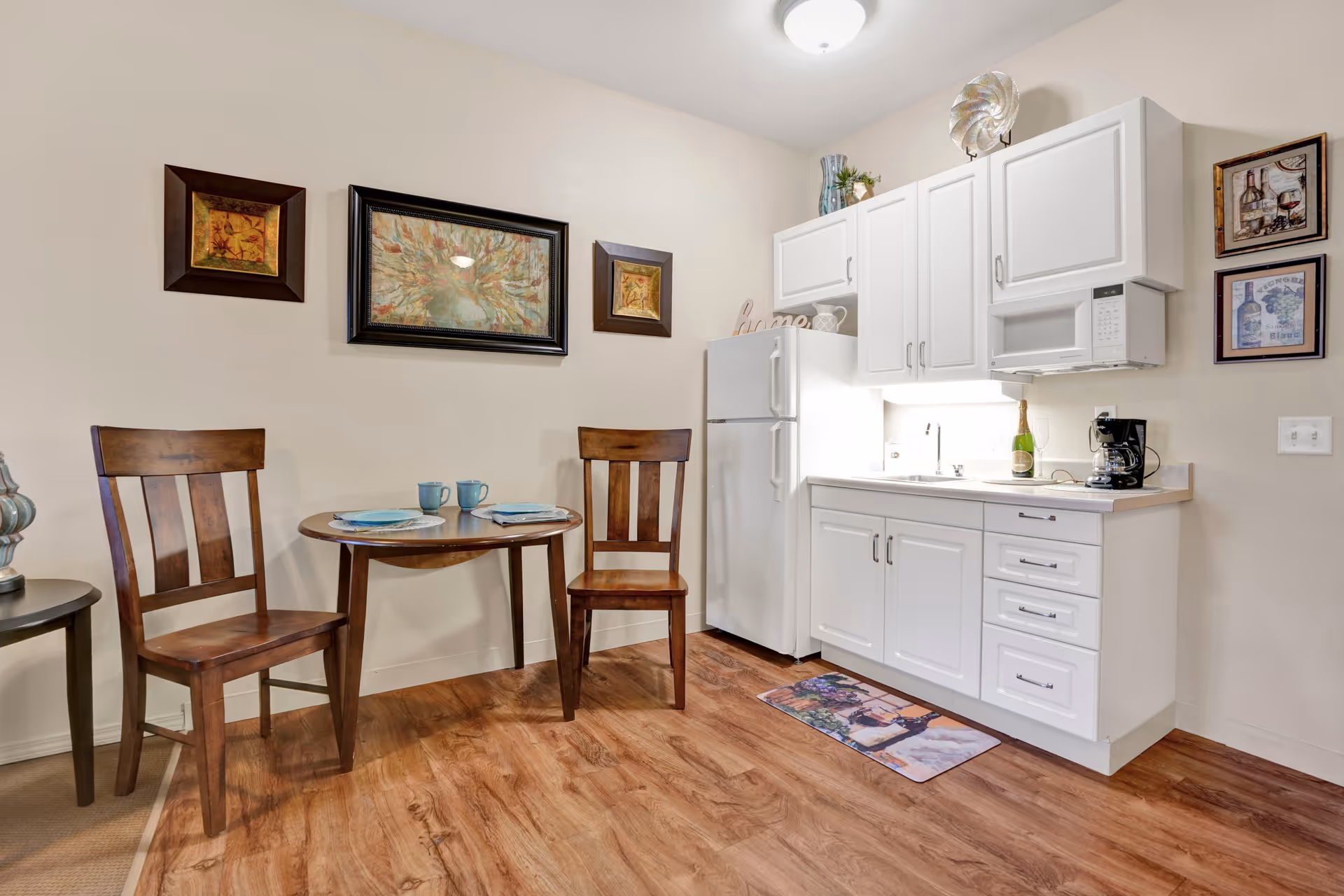 Small kitchenette and dining area with white cabinets and refrigerator, a sink and microwave, a round table set with two wooden chairs, and framed wall art.