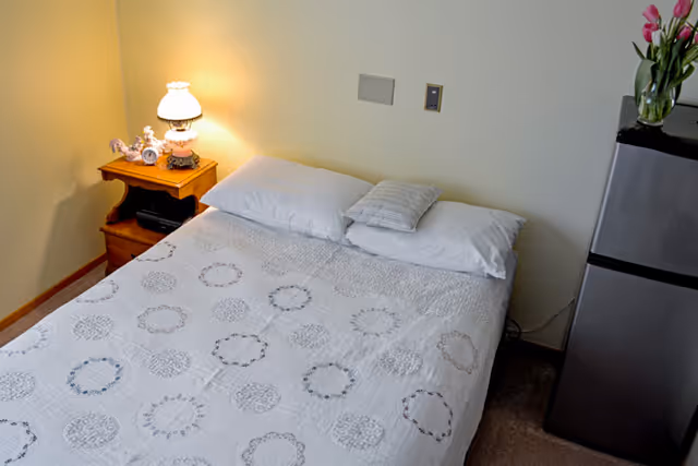 A neatly made bed with a white quilt featuring circular patterns, two pillows, and a small decorative pillow. To the left of the bed is a wooden nightstand with a vintage-style lamp and decorative items. To the right of the bed is a small refrigerator with a vase of pink flowers on top. The room has light-colored walls and carpeted floor.