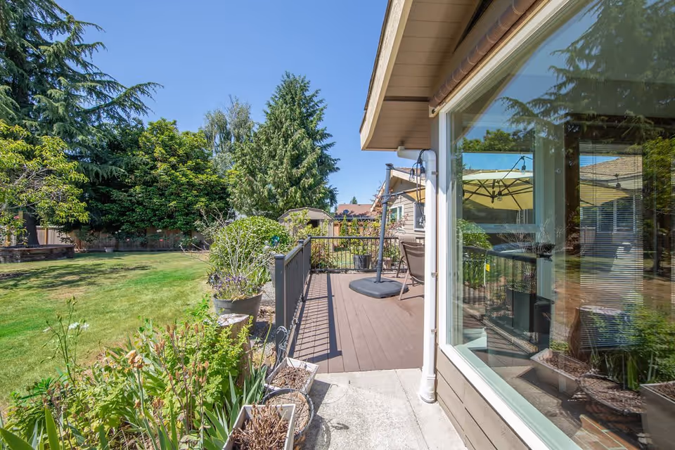 View of a backyard garden area with green grass, various plants, and trees. There is a wooden deck attached to a house with outdoor furniture and a large umbrella. The house has large windows reflecting the outdoor scene.