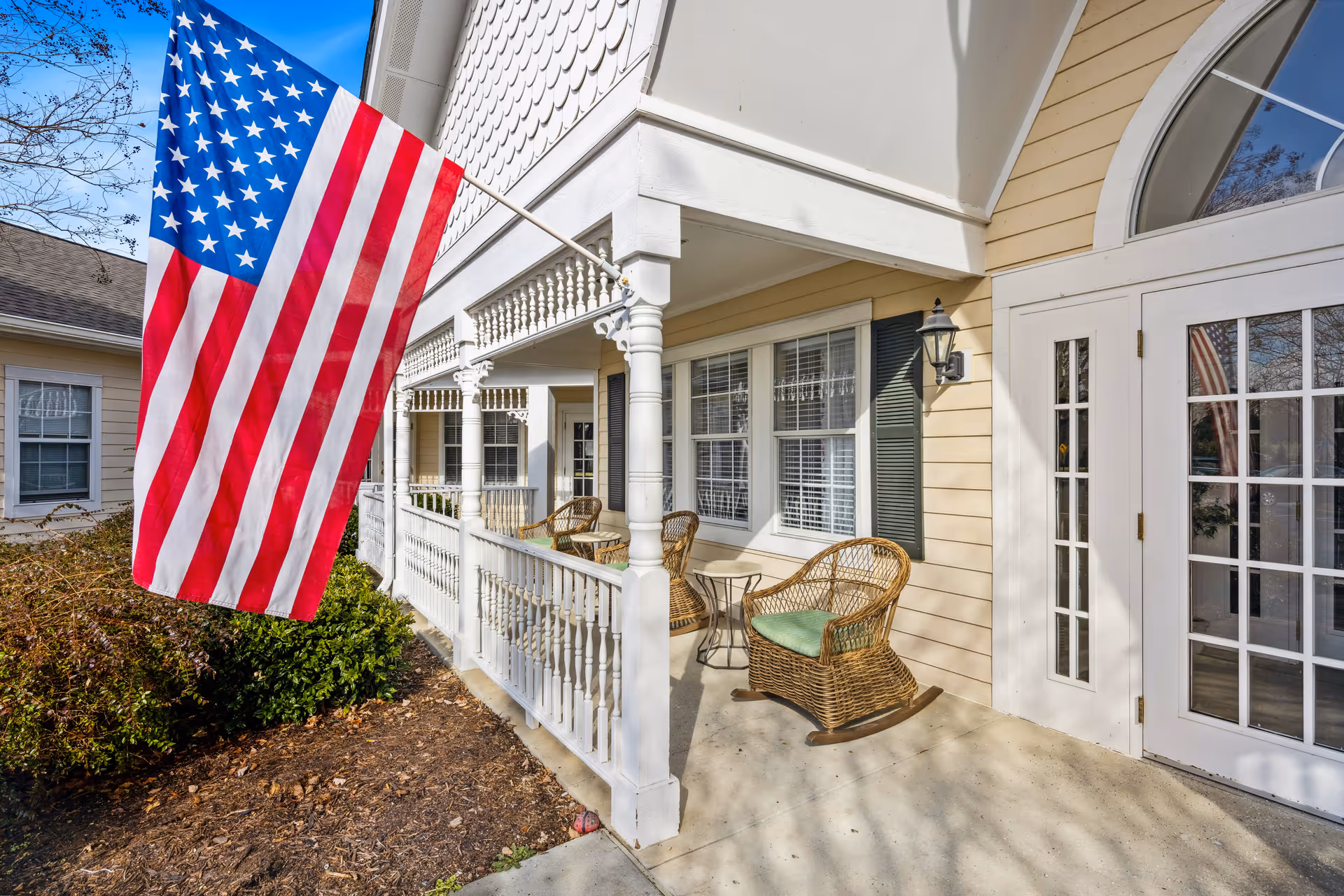 Front porch area of a senior living facility with an American flag hanging on a pole. The porch has white railings and columns, wicker rocking chairs with green cushions, a small table, and large windows with white blinds. The building exterior is light yellow with white trim and a glass door with multiple panes.