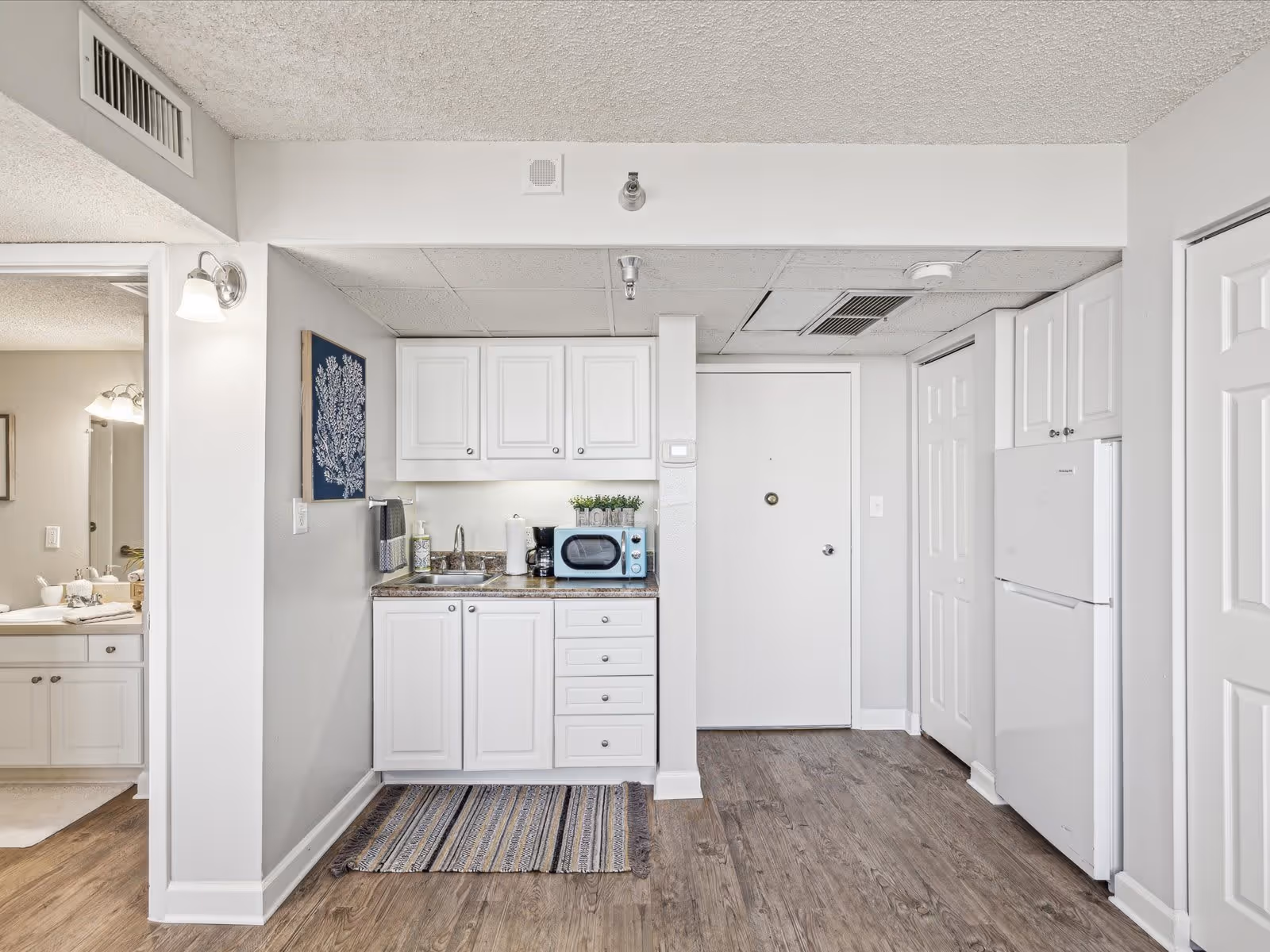 Studio kitchenette with white cabinets, sink, microwave, and refrigerator beside an entry door, with a bathroom visible to the left.