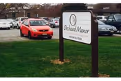 A lawn-mounted sign that reads 'Orchard Manor' with parked cars and a parking lot behind it.