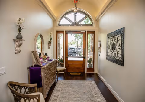 A bright entryway with a wooden front door featuring glass panels and an arched window above. The space has light-colored walls, dark wooden flooring, a patterned area rug, a decorative wooden console table with a mirror above it, a chair, and wall-mounted shelves with plants and decor. A decorative metal wall art piece is hung on the right wall.