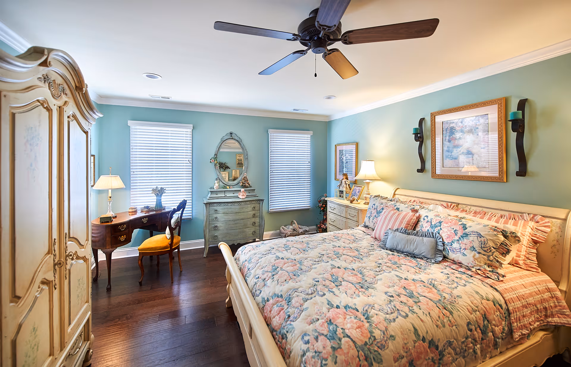 A cozy bedroom with a large bed covered in floral bedding and multiple pillows. The room features light blue walls, two windows with white blinds, a ceiling fan, a vintage-style wooden desk with a yellow cushioned chair, a dresser with an oval mirror, and a nightstand with a lamp and framed photos. There is also a large ornate wardrobe on the left side.