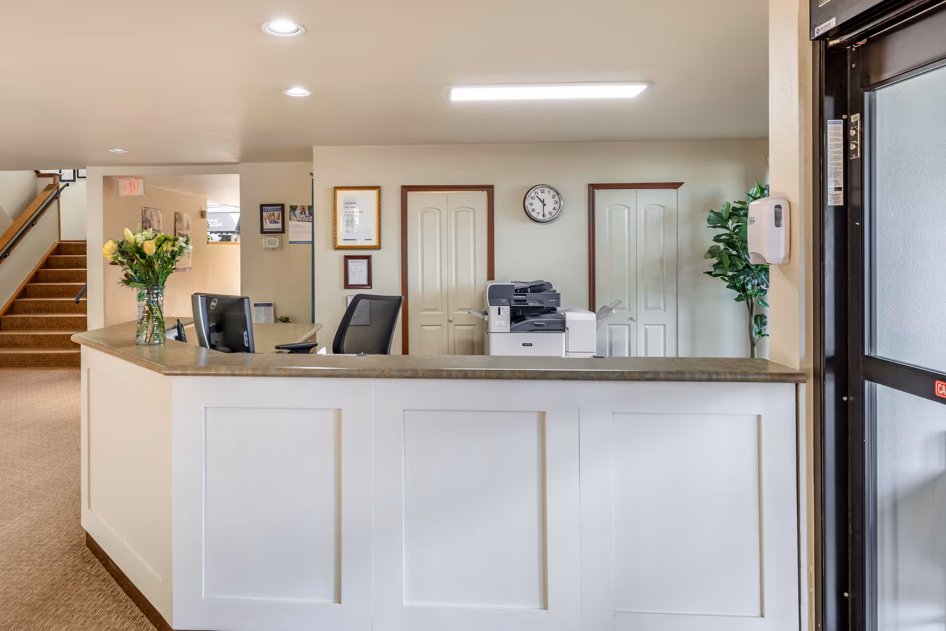 Reception desk area with a computer, office chair, printer, and a vase of yellow flowers. Behind the desk are two closed doors, a clock on the wall, framed certificates, and a potted plant. To the left, a staircase leads upstairs, and to the right, a glass door with a hand sanitizer dispenser is visible.