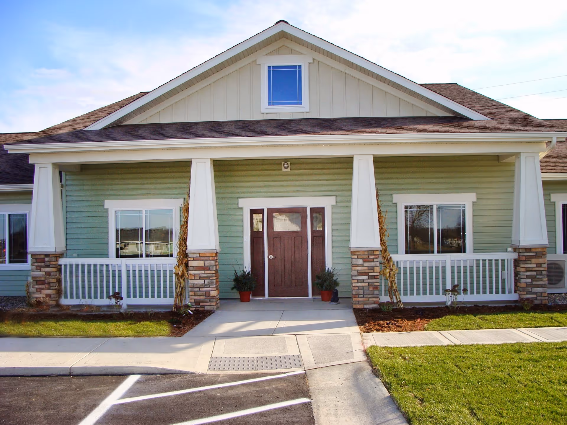 Front exterior view of a single-story building with green siding, white trim, and a brown double door entrance. The building has a covered porch supported by white columns with stone bases, two windows on either side of the door, and a small window in the gable above. There are potted plants near the entrance and a paved walkway leading to the door.