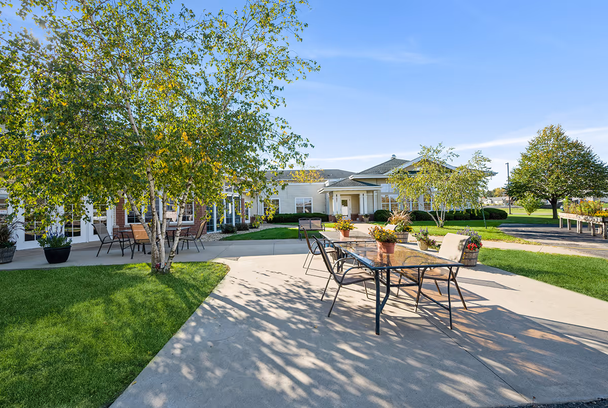 Outdoor patio area at Trustwell Living at Eagle Pointe Place featuring a glass-top table with chairs and potted plants on a concrete surface, surrounded by green grass, trees, and a building in the background under a clear blue sky.