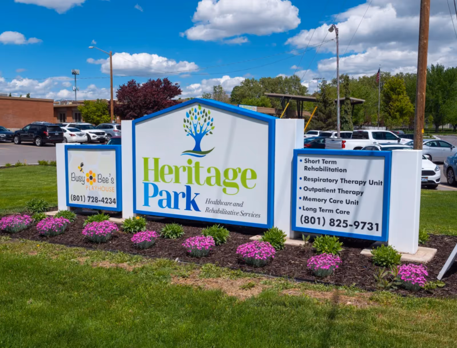 Outdoor view of a sign for Heritage Park Healthcare and Rehabilitative Services with additional signs for Busy Bee's Playhouse and a list of services including short term rehabilitation, respiratory therapy unit, outpatient therapy, memory care unit, and long term care. The sign is surrounded by green grass and purple flowers, with parked cars and trees in the background under a partly cloudy sky.