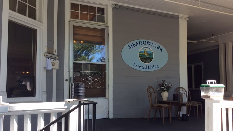 Front porch of Meadowlark Assisted Living facility with a door, two chairs, a small table with a flower vase, and a sign on the wall displaying the facility name and logo.