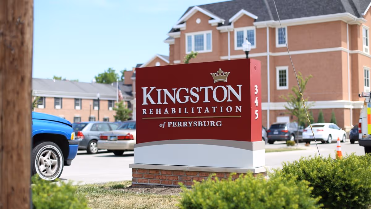 Outdoor view of a red and white sign for Kingston Rehabilitation of Perrysburg with the number 345 on the side, set in front of a parking lot with several cars and a multi-story brick building in the background.
