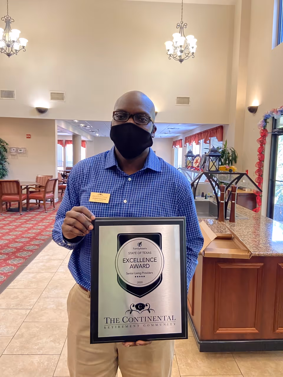A man wearing glasses and a black face mask stands inside a retirement community lobby holding a framed Excellence Award plaque from the State of Texas for senior living providers. The lobby features tiled floors, red patterned carpet, wooden chairs and tables, chandeliers, and a counter with decorative items.
