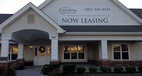 Exterior front view of a single-story building with a covered entrance. The building has beige siding with white trim and brick accents. Above the entrance, there is a large sign that reads 'Heartsong Memory Care' with a phone number and the words 'Residential and Adult Day Health Care NOW LEASING'. There are shrubs and small plants in front of the building.