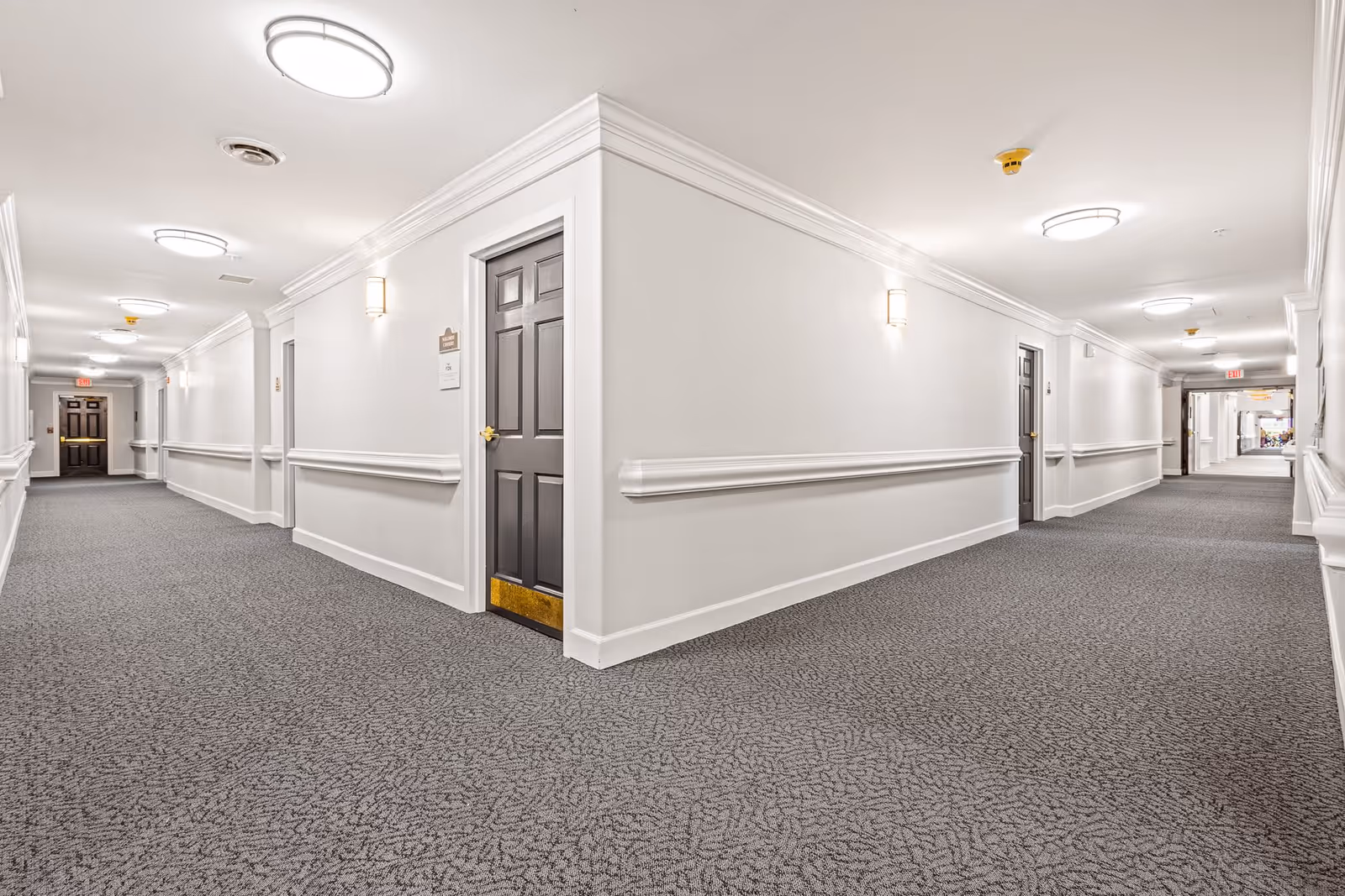 A clean, well-lit hallway in a senior living facility with gray carpet, white walls, and multiple closed dark-colored doors on both sides. Ceiling lights and wall sconces illuminate the corridor, which has handrails along the walls and an exit sign visible at the far end.