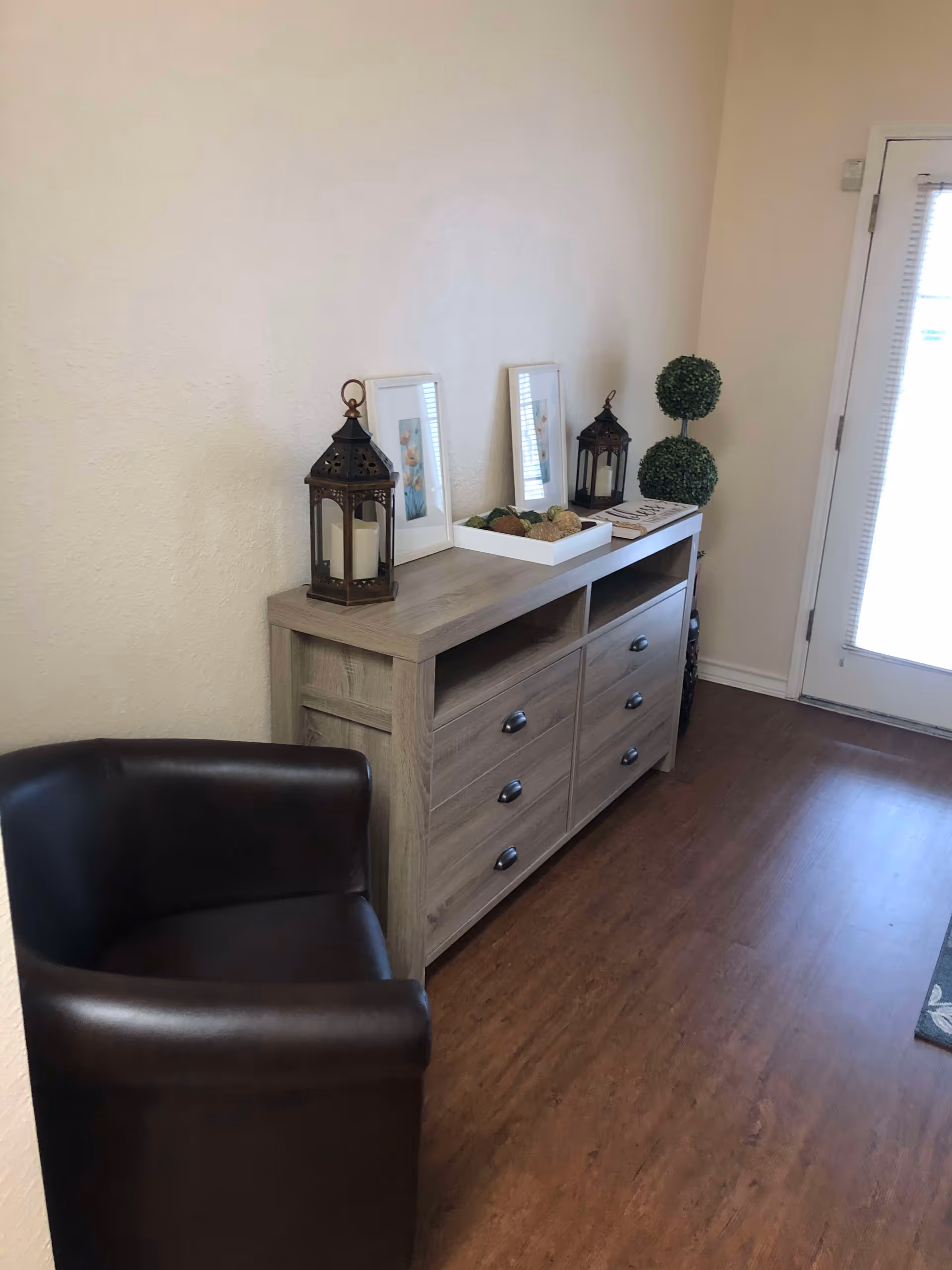 A corner of a room with a wooden dresser featuring six drawers and two open shelves. On top of the dresser are two decorative lanterns with candles, two framed pictures, a white tray with decorative balls, and a small artificial topiary plant. To the left of the dresser is a dark brown leather armchair. The floor is wooden, and there is a door with a window and blinds on the right side.