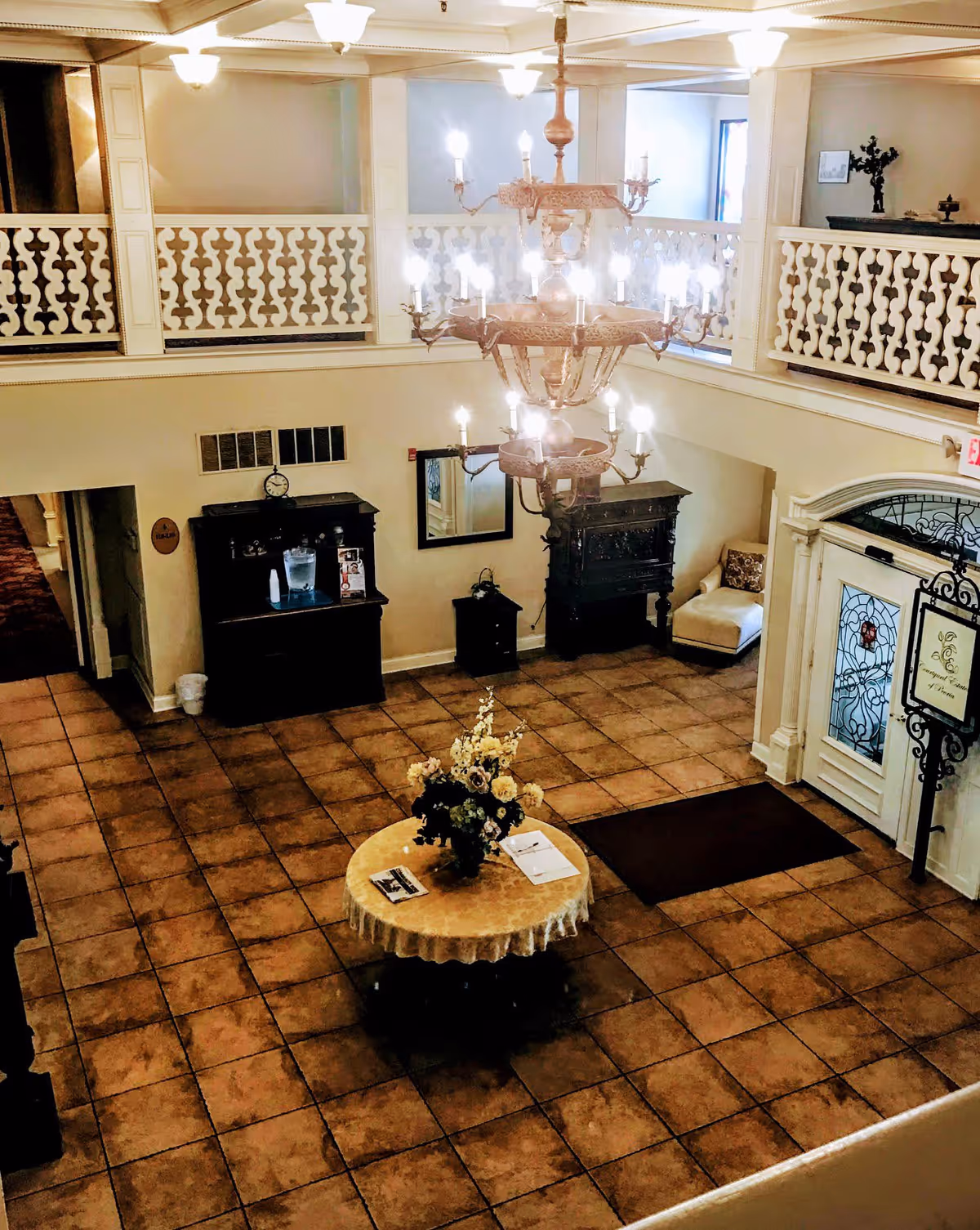 View of a spacious, elegant lobby area with a large chandelier hanging from the ceiling. The floor is tiled with a round table covered with a lace tablecloth and a floral arrangement in the center. There is a dark wooden cabinet with a water dispenser and a clock on the wall, a small seating area with a cushioned chair, and a decorative fireplace. The entrance door has decorative glass panels and a sign next to it. The upper level has a white railing with a decorative pattern.