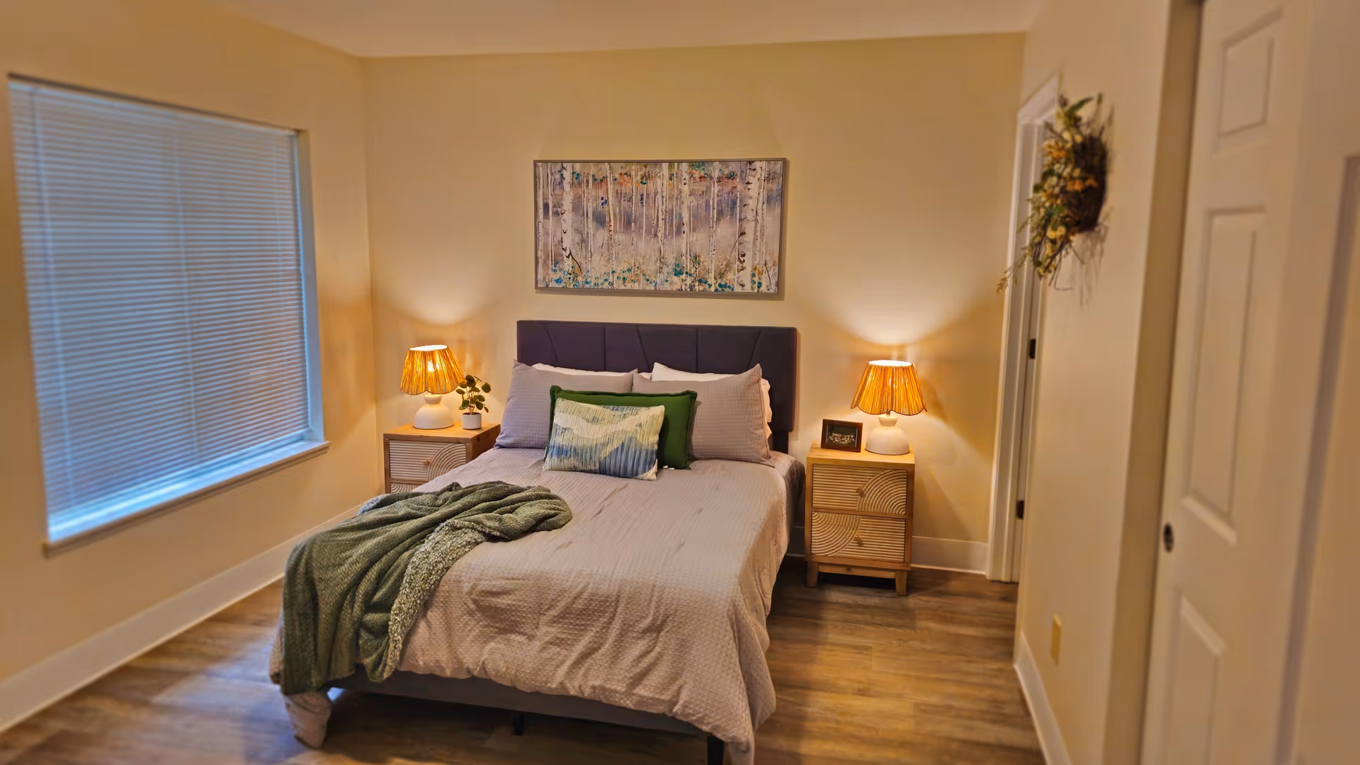 Well-lit bedroom with a made bed, two bedside tables with lamps, a window with blinds, and wall art above the headboard.