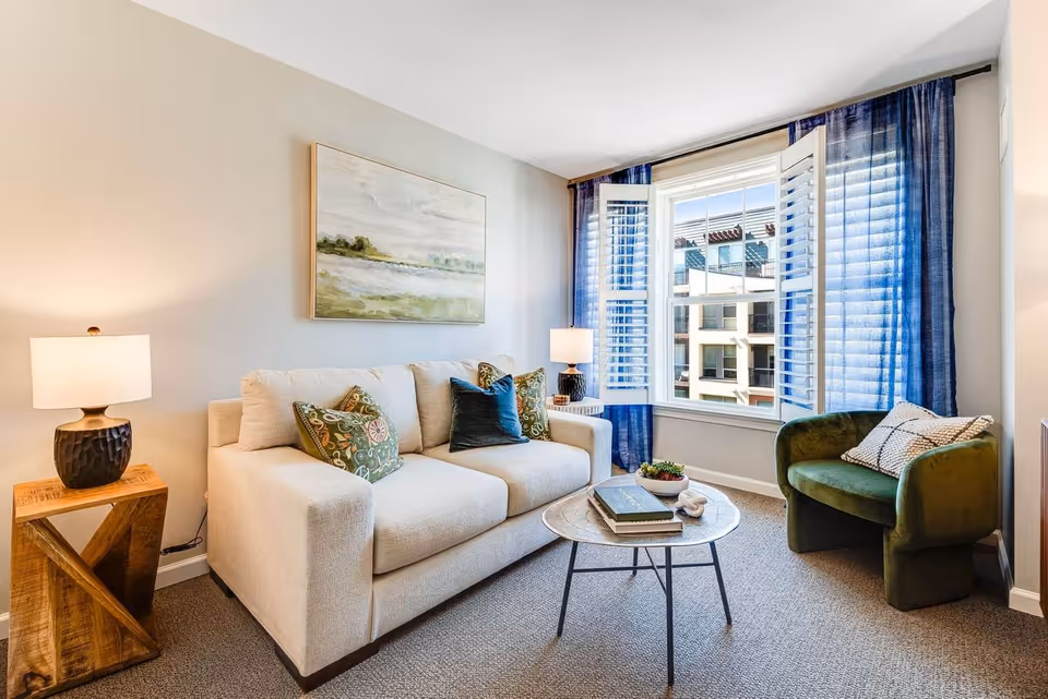 A cozy living room with a beige sofa adorned with patterned and solid pillows, a round coffee table with books and a small plant, a green armchair with a cushion, two table lamps on wooden side tables, and a large window with blue curtains and white shutters letting in natural light.