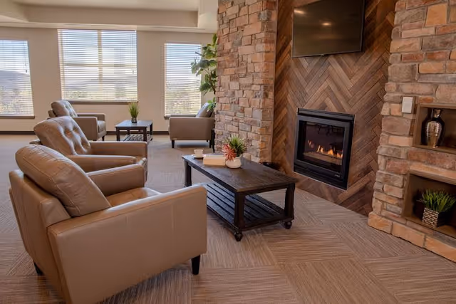 A cozy living room area with beige leather armchairs arranged around a dark wooden coffee table. The room features a stone fireplace with a fire burning and a mounted flat-screen TV above it. Large windows with blinds allow natural light to fill the space, and there are small plants on the coffee table and in the corner near the windows.