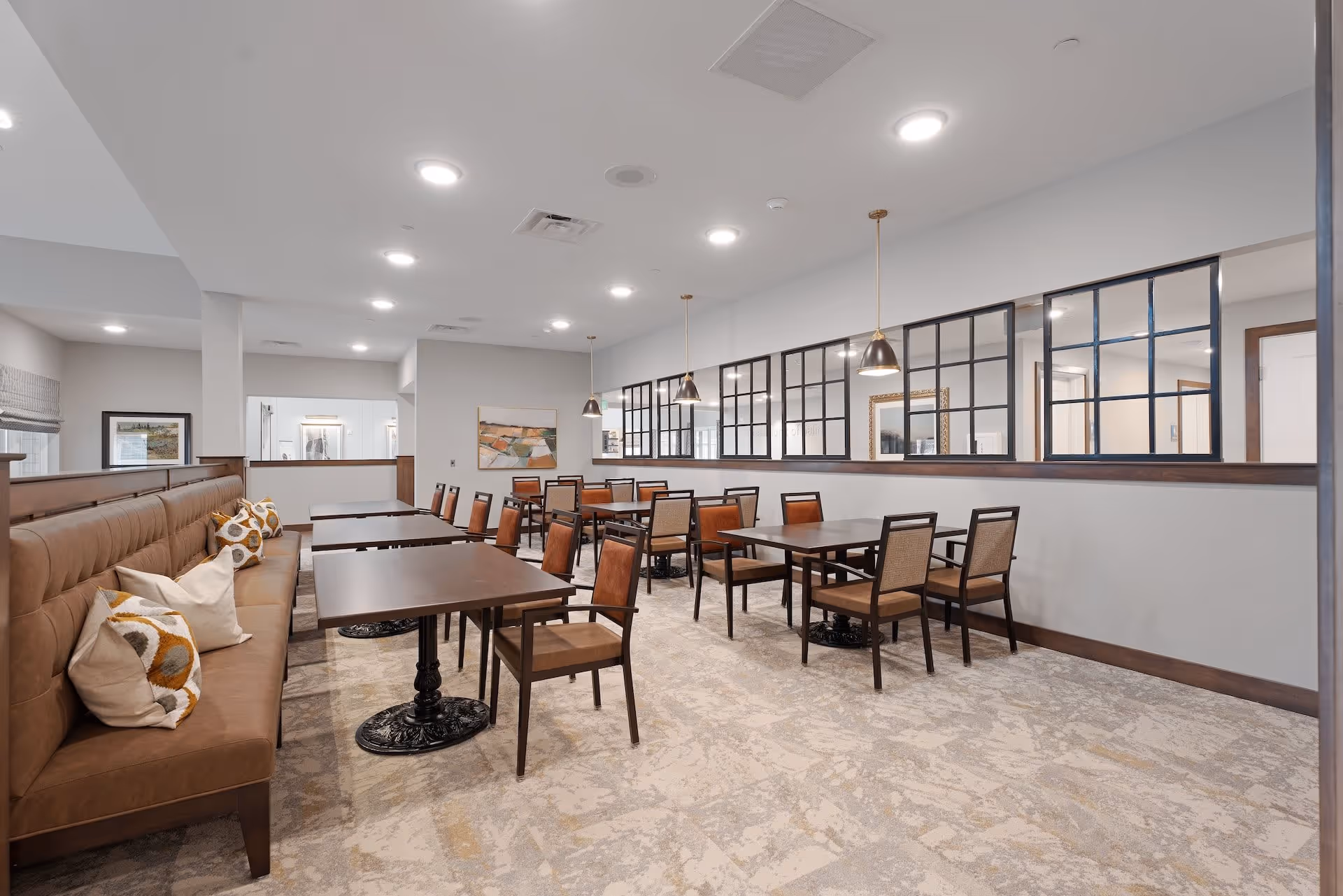 Spacious dining room with multiple wooden tables and chairs, banquette seating with pillows, pendant lights, and interior grid windows.