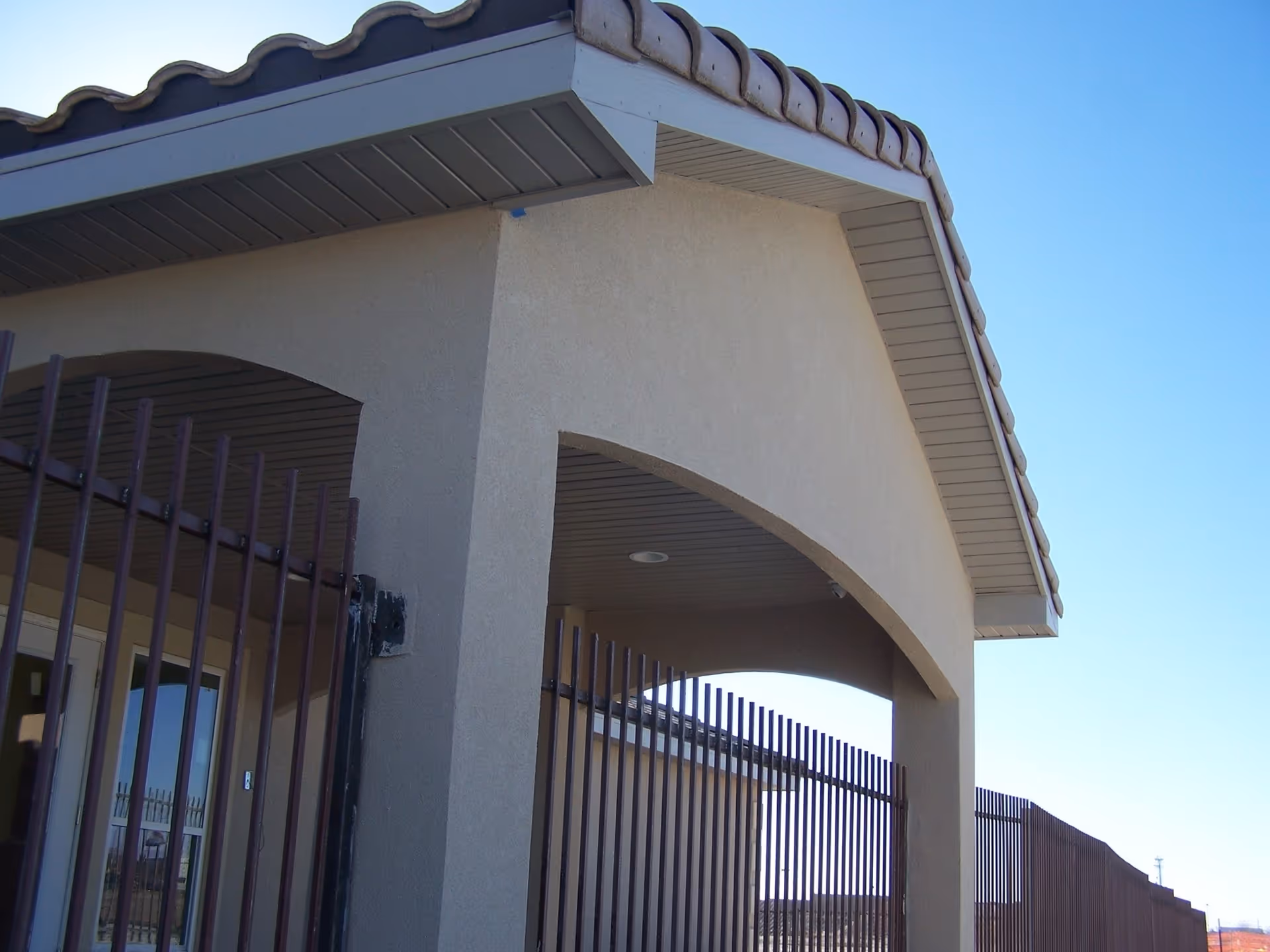 Exterior view of a building corner with a beige stucco finish and a tiled roof. There is a metal fence with vertical bars surrounding the building, and the sky is clear and blue.