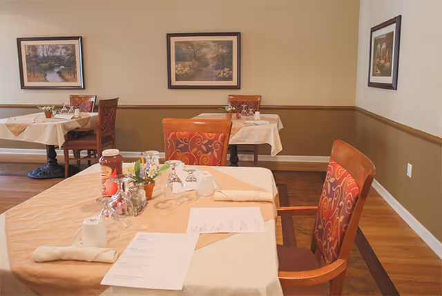 A dining room with tables covered in beige tablecloths, set with menus, napkins, glassware, and condiments. The chairs have red and orange patterned upholstery. The walls are painted beige and brown with framed landscape paintings hanging on them. The floor is wooden.