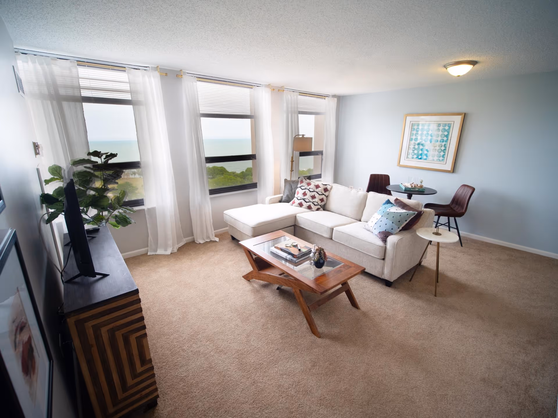A bright living room with large windows covered by sheer white curtains, offering a view of greenery and water outside. The room features a light beige sectional sofa with decorative pillows, a wooden coffee table with books and decor, a small round side table, a TV on a wooden stand, and a small round dining table with two chairs. The walls are painted light blue and there is a framed artwork hanging on the wall.