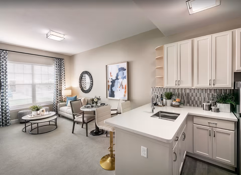 A bright and modern living area with a small kitchen. The kitchen features white cabinets, a white countertop with a sink, and a gray patterned backsplash. Adjacent to the kitchen is a cozy living room with a beige sofa, a round coffee table, a small dining table with chairs, and large windows with patterned curtains letting in natural light. The walls are decorated with a round mirror and an abstract painting.