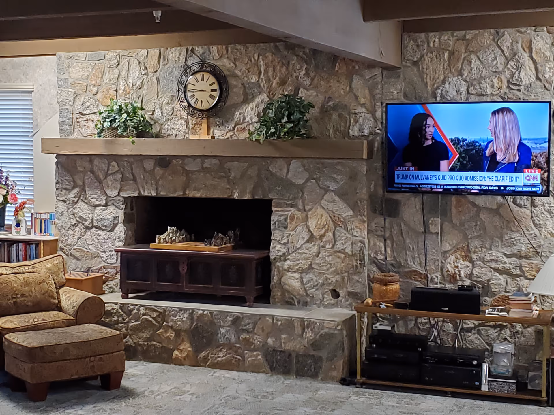 Cozy living room with a stone fireplace featuring a wooden mantel decorated with two green plants and a round clock. To the left, there is a comfortable patterned armchair with an ottoman, a bookshelf with books and flowers, and a window with blinds. On the right side, a flat-screen TV mounted on the stone wall displays a CNN news segment with two women talking.