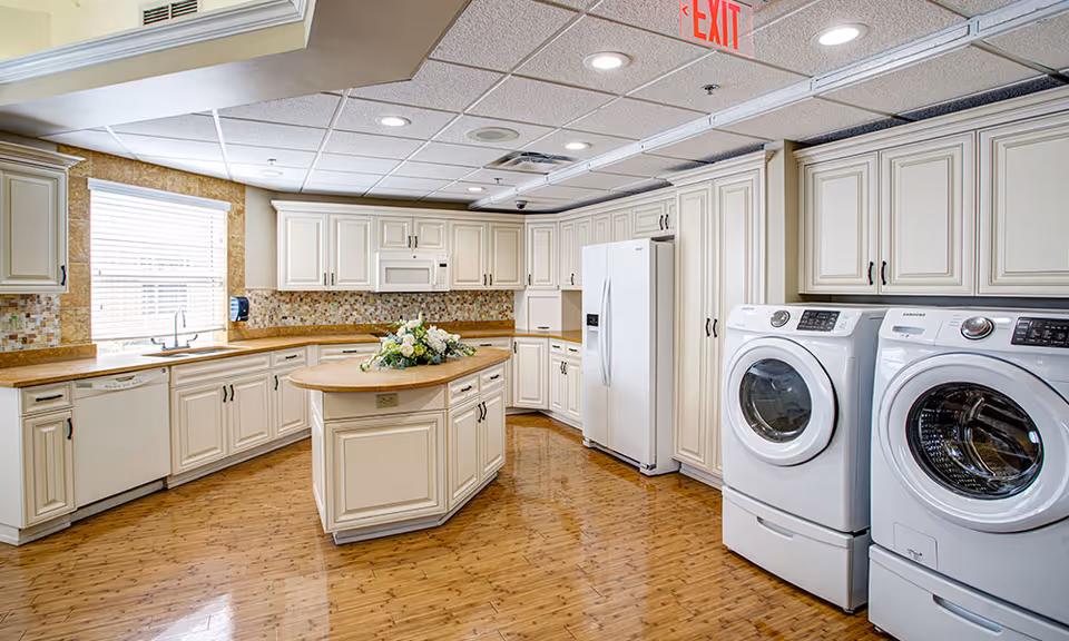 Bright communal kitchen with white cabinetry, a central island, refrigerator, and front-loading washer and dryer along the right wall.
