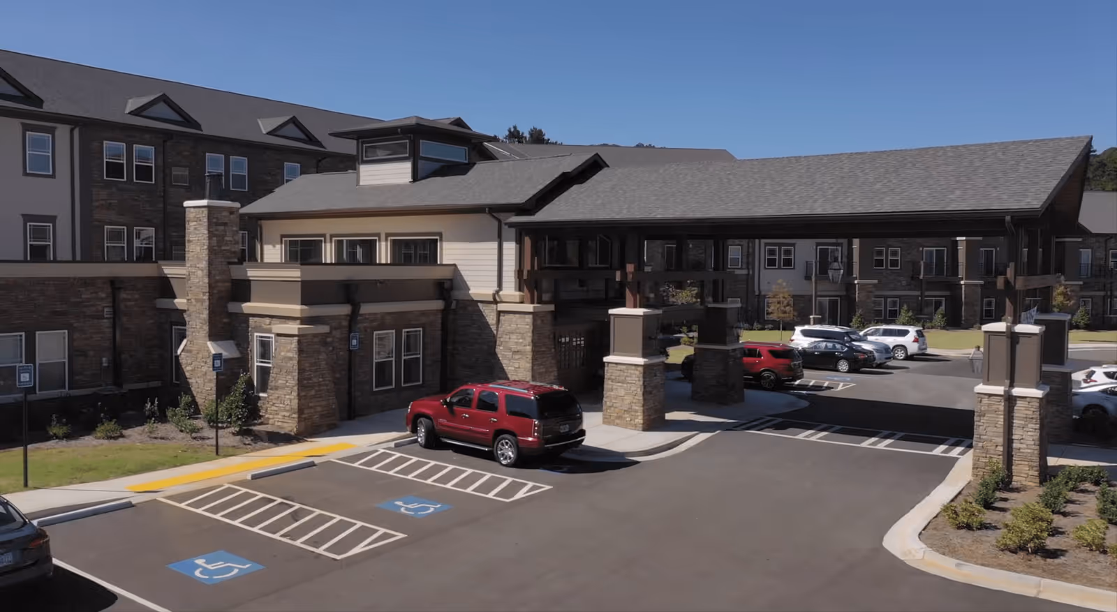 Front exterior of a senior living facility showing a covered entrance with stone pillars, adjoining parking lot, handicap spaces, and several parked cars.