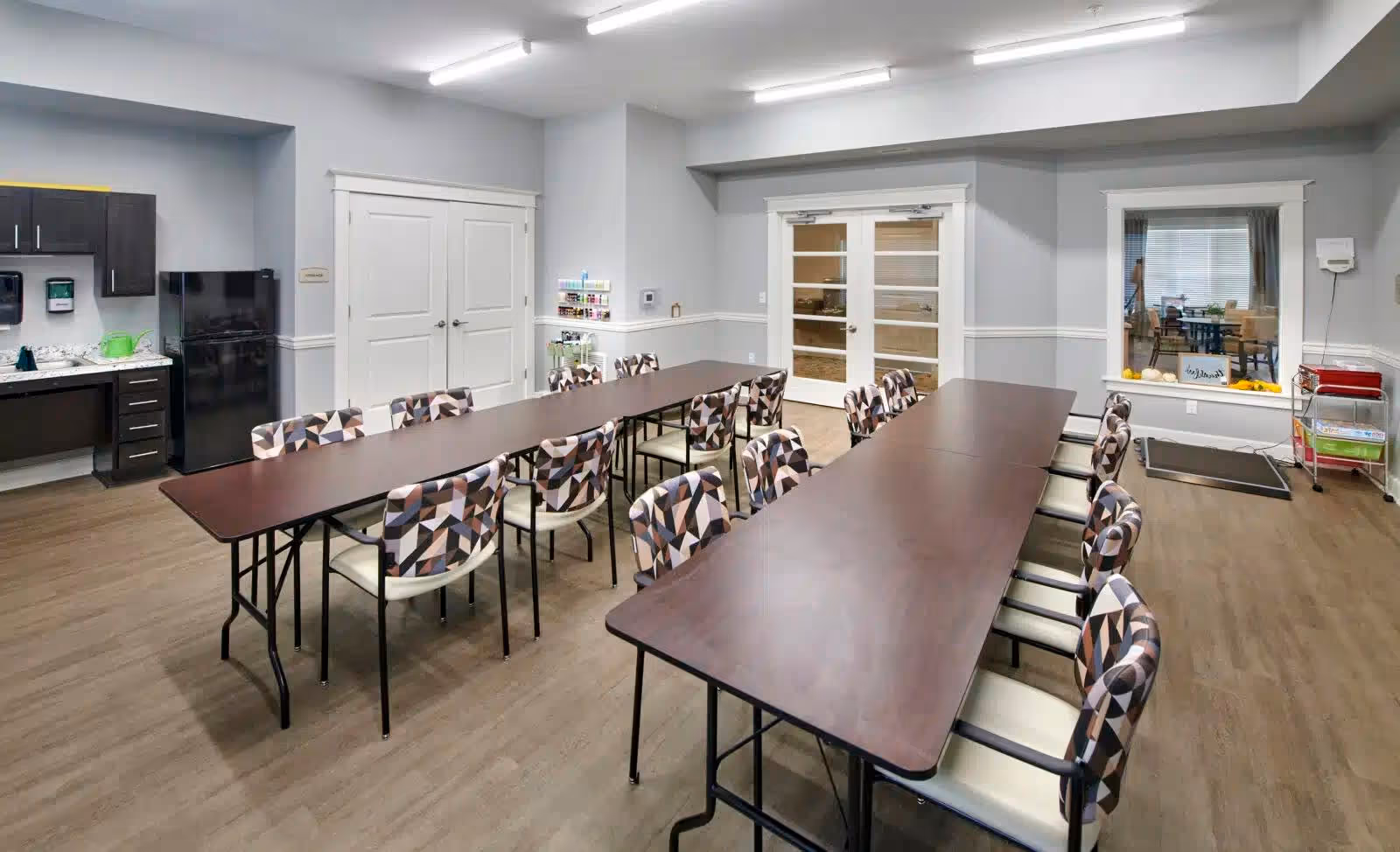 A well-lit room with two long rectangular tables arranged in a U-shape, surrounded by patterned chairs. The room has light gray walls, wood flooring, and a small kitchenette area with a black refrigerator and dark cabinets. There are double white doors and a window looking into another room, with a small cart and some supplies near the window.
