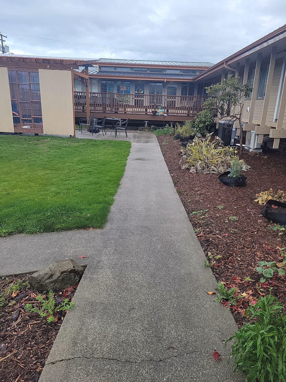 Outdoor pathway leading to a wooden deck area with chairs and tables, surrounded by green grass on the left and mulched garden beds with plants on the right, under a cloudy sky.
