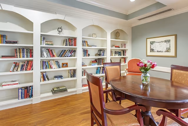 A cozy room with a wooden round table surrounded by four chairs with patterned cushions. Behind the table, there are built-in white bookshelves filled with books and decorative items. A vase with flowers is placed on the table, and a framed picture hangs on the wall. The room has wooden flooring and soft lighting.
