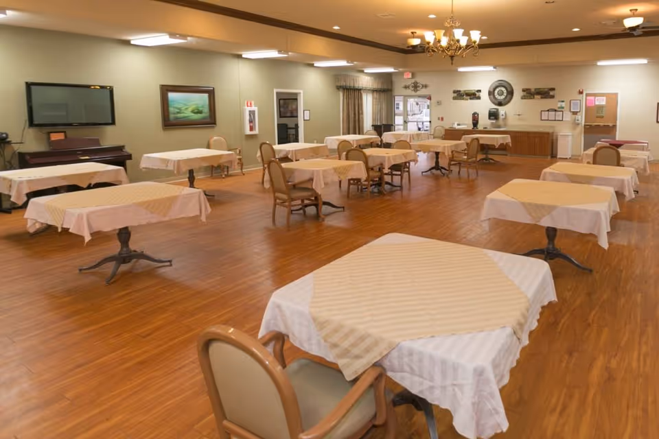 A spacious dining room with multiple tables covered in beige and white tablecloths, each surrounded by chairs. The room has wooden flooring, a chandelier, a wall-mounted TV, framed artwork, and a counter area with coffee dispensers and a clock on the wall.