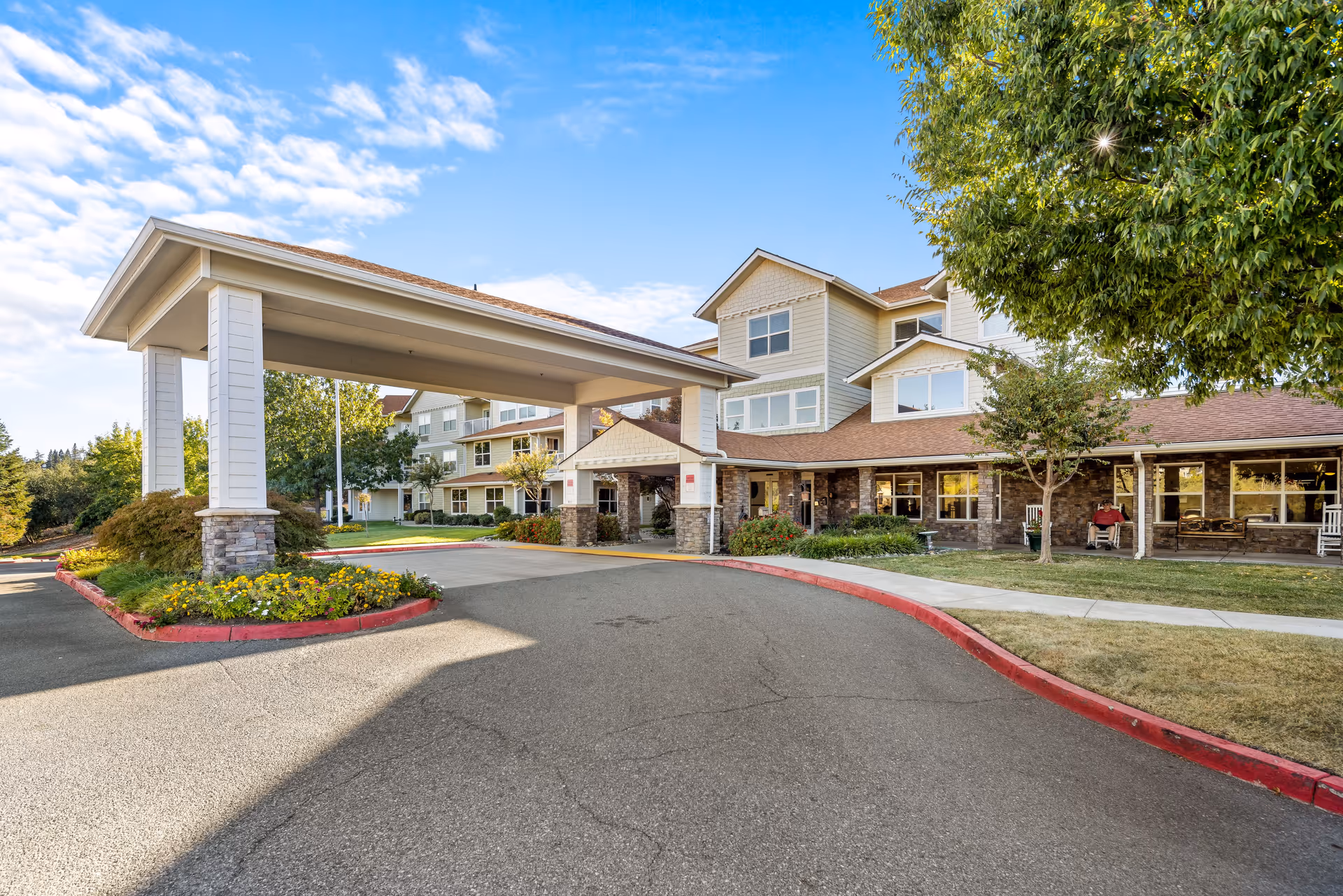 Exterior view of a senior living facility with a covered entrance driveway, landscaped flower beds, trees, and a multi-story building with large windows under a partly cloudy sky.