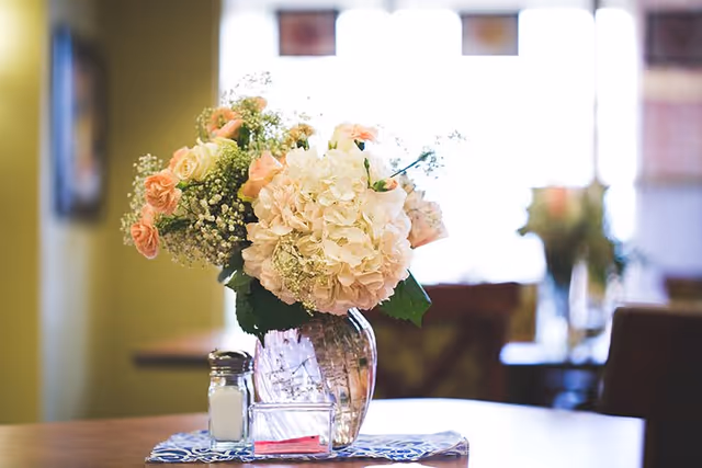A glass vase of pink and white flowers serving as a centerpiece on a table in a softly lit dining area.