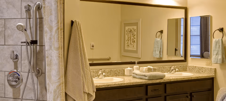 A bathroom with a double sink vanity featuring a granite countertop, two mirrors above the sinks, and neatly folded towels. To the left is a tiled shower area with a handheld showerhead and a grab bar. Light-colored walls and soft lighting create a warm atmosphere.