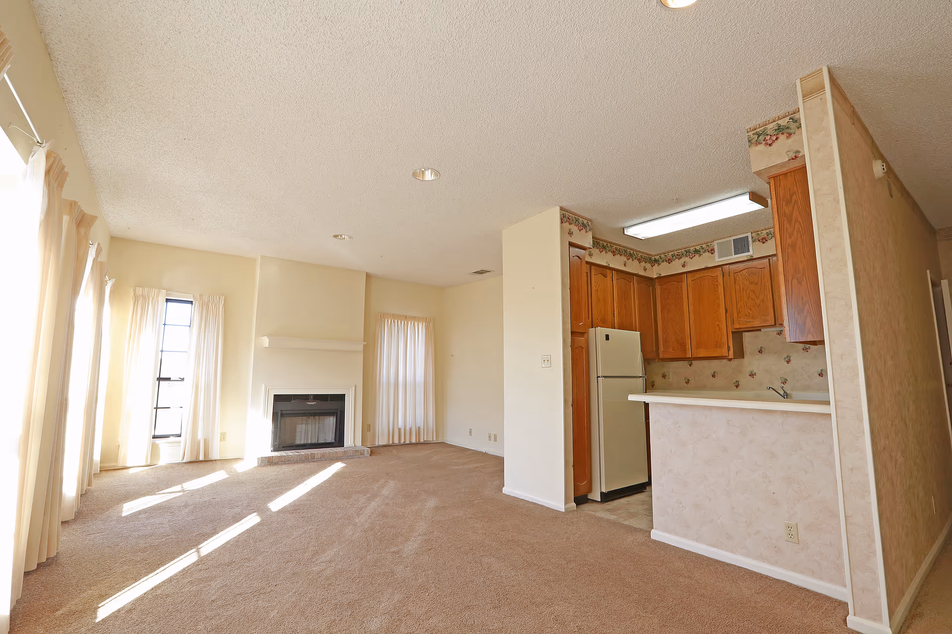 A bright living room with beige carpet, cream-colored walls, and large windows with sheer curtains allowing natural light to fill the space. There is a fireplace centered on one wall. Adjacent to the living room is a kitchen area with wooden cabinets, a white refrigerator, and a countertop with a small breakfast bar.