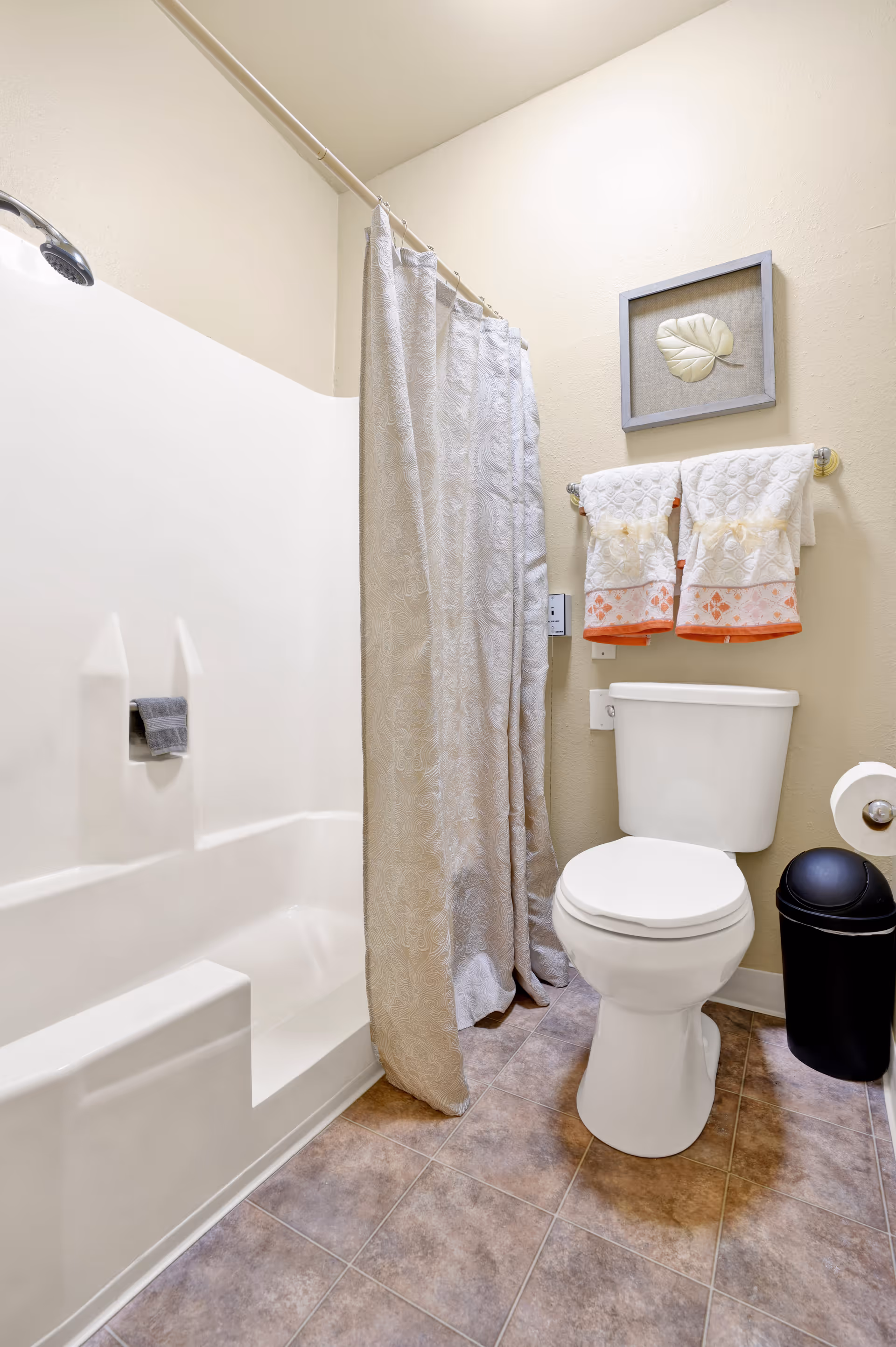 A clean bathroom featuring a white bathtub with a showerhead and a beige patterned shower curtain. Next to the bathtub is a white toilet with a black trash can beside it. Above the toilet, there are two white and orange decorative towels hanging on a towel rack, and a framed leaf artwork on the wall. The floor is covered with brownish tiles.